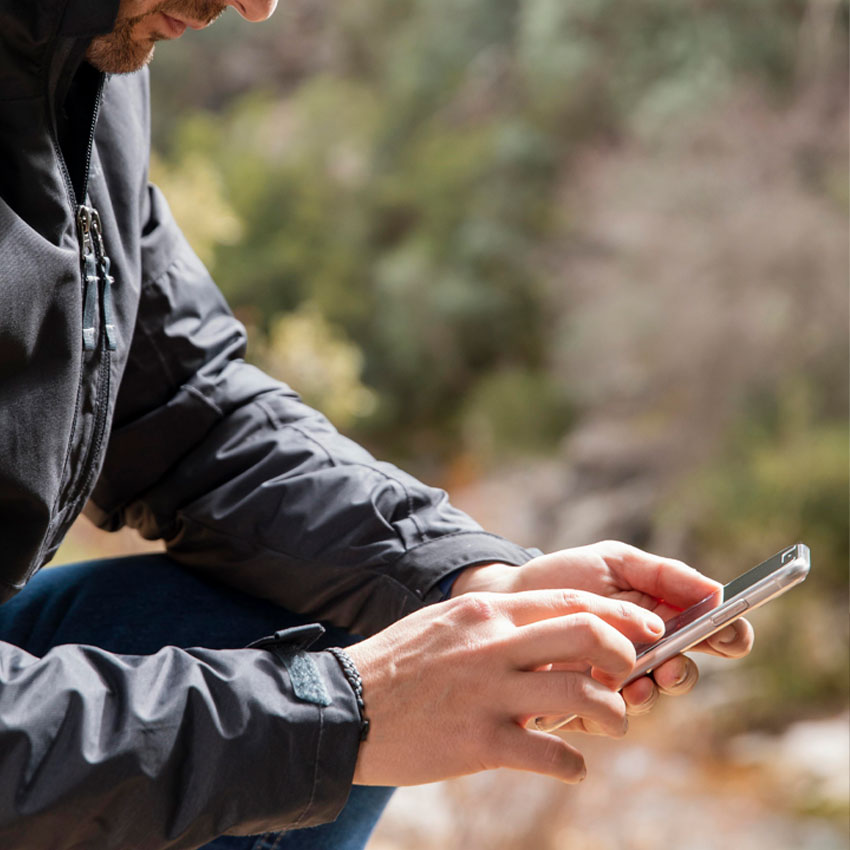 A person wearing a dark jacket is outdoors, using a smartphone with both hands. The background is blurred, showing greenery and natural surroundings.