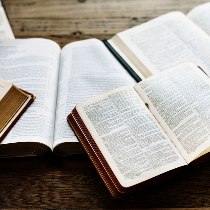 Several open books with visible text lie spread out on a wooden table, overlapping each other. The pages are filled with dense, closely printed text, suggesting study or research in progress.